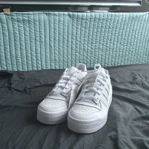 Vita sneakers från Adidas - Snygga vita sneakers från Adidas med klassiska tre ränder på sidan. Skorna har en robust sula och snörning framtill. Perfekta för en stilren look. Helt oanvända!!