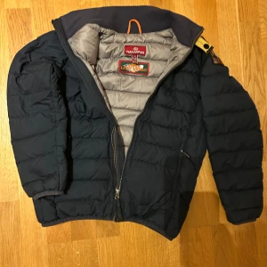 Mörkblå dunjacka från Parajumpers strl 152 - Säljer en snygg mörkblå dunjacka från Parajumpers med dragkedja och hög krage. Jackan har en praktisk ficka med dragkedja på sidan och en ikonisk patch på ärmen. Nypris 3995:-