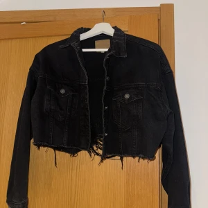 Svart croppad jeansjacka - Snygg svart croppad jeansjacka med slitna detaljer och knappar framtill. Perfekt för en cool och avslappnad stil. Jackan har långa ärmar och två bröstfickor med lock.