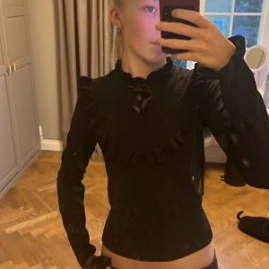 Svart tröja med volang från Holly & Whyte - Snygg svart tröja från Holly & Whyte med volangdetaljer runt halsen och en liten knytning framtill. Perfekt för en stilren look.