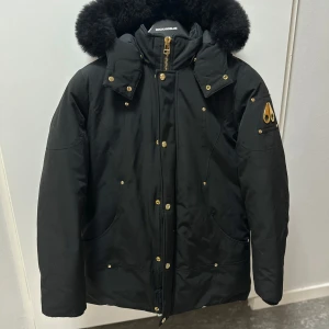Svart vinterjacka från Moose Knuckles - Snygg svart vinterjacka från Moose Knuckles med pälskantad huva och guldiga detaljer. Jackan har dragkedja och knappar framtill samt ett broderat märke på ärmen. Perfekt för kalla dagar!