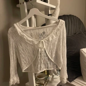 Vit spetsbolero - Säljer en söt vit bolerotopp i spets med knytning framtill. Toppen har långa ärmar och en fin volangdetalj vid ärmslut och halsringning. 