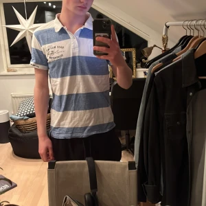 Blåvit randig pikétröja från Race Marine - Säljer en stilren blåvit randig pikétröja från Race Marine. Tröjan har en klassisk krage med knappar och tryck på bröstet. Perfekt för en avslappnad stil.