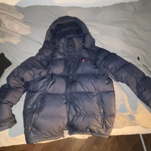 Mörkblå dunjacka från Polo Ralph Lauren - Säljer en snygg mörkblå dunjacka från Polo Ralph Lauren. Jackan har en huva och dragkedja framtill samt två praktiska fickor med dragkedjor. Perfekt för kyliga dagar!