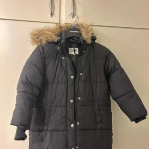 Svart dunjacka med päls från KappAhl - Säljer en svart dunjacka från KappAhl med avtagbar huva och pälsdetaljer. Jackan har knappar och dragkedja framtill samt långa ärmar. Perfekt för kalla vinterdagar.