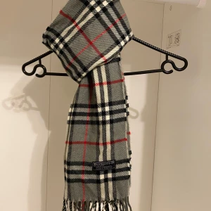 Burberry - Säljer nu denna Snygga halsduk från Burberry i färgen grå. Tillverkad i mjukt lammull med klassiskt fransad kant. Perfekt för att hålla dig varm och stilren under kyliga dagar. Hör av er vid funderingar☺️