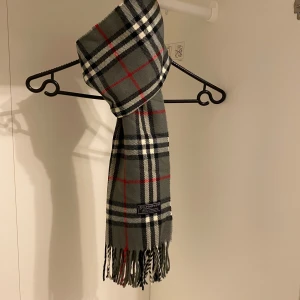 Burberry - Säljer nu denna Snygga halsduk från Burberry i färgen grå. Tillverkad i mjukt lammull med klassiskt fransad kant. Perfekt för att hålla dig varm och stilren under kyliga dagar. Hör av er vid funderingar☺️