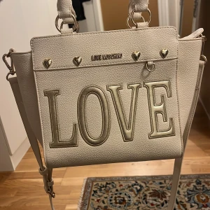 Vit axelväska från Love Moschino - Snygg vit axelväska från Love Moschino med stort 'LOVE' tryck på framsidan och hjärtformade detaljer. Väskan har en praktisk dragkedja och är tillverkad i skinnmaterial. Perfekt för att ge en stilfull touch till din outfit.