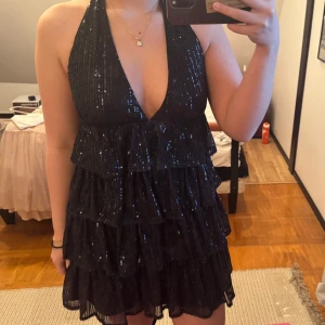 Svart glittrig klänning - Snygg svart klänning med glittriga detaljer och djup v-ringning. Klänningen har volanger och är ärmlös med halterneck-design. Perfekt för nyår eller en fest! 
