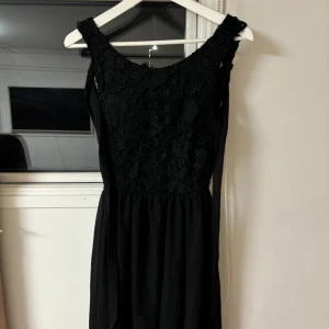 Svart klänning från Model Behaviour - Elegant svart klänning med spetsdetaljer och öppen rygg från Model Behaviour. Klänningen har en knytning i ryggen och är ärmlös, vilket ger en stilren och sofistikerad look. Perfekt för speciella tillfällen.