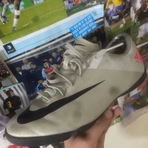 Nike Mercurial fotbollsskor - Snygga Nike Mercurial fotbollsskor i silver med svarta och röda detaljer. Skorna har en smidig snörning och en slitstark sula, perfekt för inomhusfotboll. Den ikoniska Nike-loggan syns tydligt på sidan. Säljer dessa för att rensa ut min fotbollsamling 