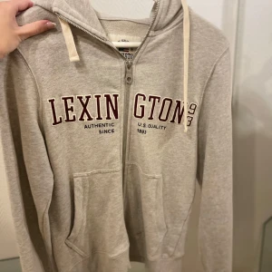 Beige hoodie från Lexington - Säljer en beige hoodie från Lexington med dragkedja och tryck framtill. Den har en justerbar huva med snören och praktiska fickor. Perfekt för en avslappnad stil.