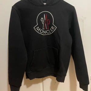 Svart hoodie från Moncler - Säljer en stilren svart hoodie från Moncler med deras ikoniska logga broderad på framsidan. Tröjan har en klassisk känguruficka och ribbade muddar vid ärmslut och nederkant. Perfekt för kyliga dagar! Pris kan diskuteras vid snabb affär