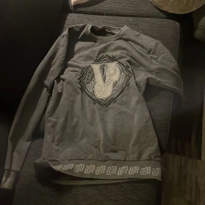  tröja  - Säljer en snygg grå tröja från Versace med ett stort V-märke på framsidan. Tröjan har långa ärmar och ett dekorativt mönster längs nederkanten. Tillverkad i Italien av bomull och polyester.