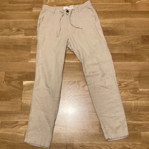 Beige linnebyxor från Selected Homme - Snygga beige linnebyxor från Selected Homme med snörning i midjan och knappstängning. Perfekta för en avslappnad stil med två fickor fram och bak. Passar bra till både vardag och lite mer uppklädda tillfällen men passar också i vardagen