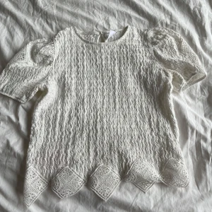 Vit spetsblus från Zara - Säljer en söt vit spetsblus från Zara med korta puffärmar och dekorativ kant nedtill. Perfekt för en somrig look eller speciella tillfällen. Blusen har ett fint mönster och en knapp i nacken för enkel påklädning.