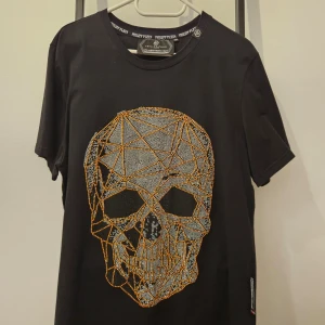 Svart t-shirt med dödskalle från Philipp Plein - Cool svart t-shirt från Philipp Plein med en stor, glittrig dödskalle i orange och silver på framsidan. 