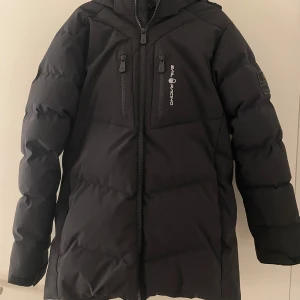 Svart dunjacka från Sail Racing (ny skick) - Snygg svart dunjacka från Sail Racing med dragkedja och huva. Jackan är tillverkad i återvunnen nylon och spandex, med fyllning av dun och fjädrar. Perfekt för kalla dagar. Köpt januari 2024 och har bara använt den några gånger. 