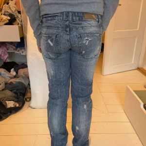 Blå jeans med slitningar - Säljer ett par blå lowwaist jeans med coola slitningar och en klassisk femficksdesign. Jeansen har en knappgylf och är i en straight passform. Perfekta för en avslappnad stil.