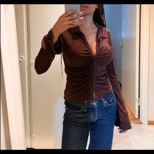 Brun, åtsittande sammetsblus - En elegant, åtsittande topp i brun sammet. Den har en smickrande rynkad detalj framtill och långa ärmar. Mjuk och bekväm, passar lika bra till avslappnade som uppklädda tillfällen. 