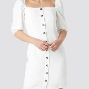 Vit kortklänning med knappar - Säljer en superfin vit kortklänning med knappar hela vägen ner. Den har puffiga trekvartsärmar och en fyrkantig urringning som ger en söt och stilren look. Perfekt för sommarens alla fester eller en dag på stan! (Pris kommer vi överens om privat🥰 )