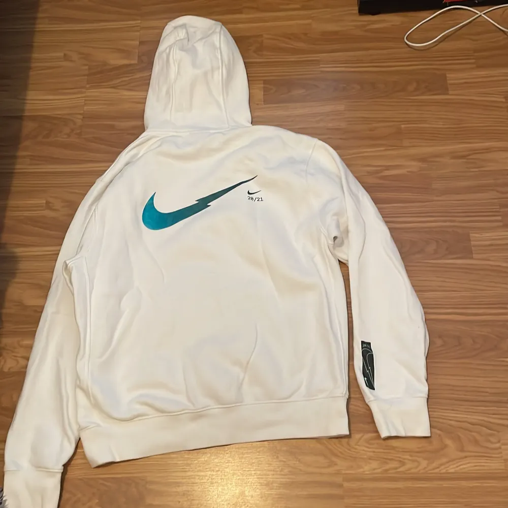 Säljer en snygg vit hoodie från Nike i storlek M. Den är i perfekt skick 10/10 med en stor Nike-logga i blått på ryggen och en mindre på framsidan. Hoodien är i standard passform och har en stor ficka framtill. Perfekt för både vardag och träning!. Hoodies.