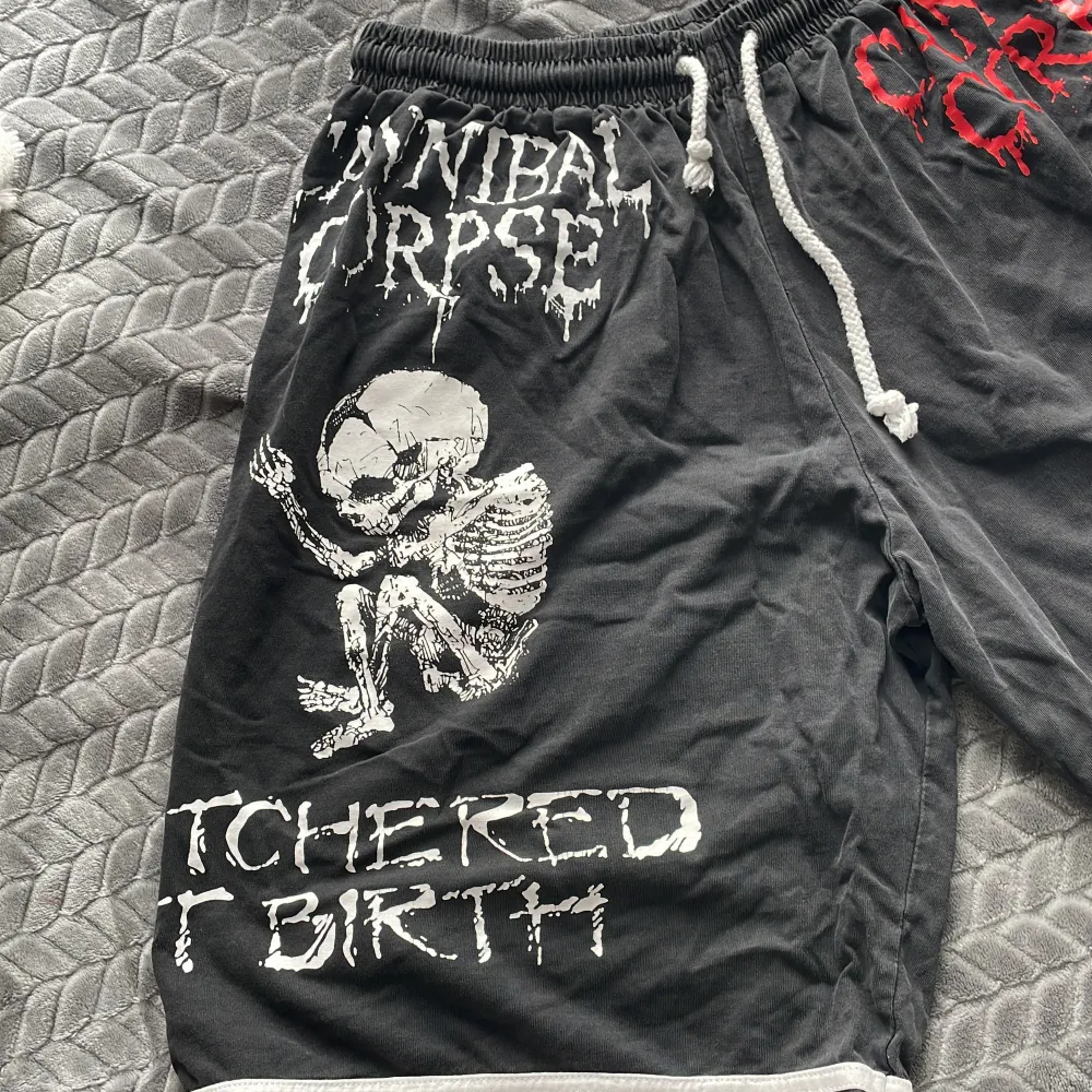 Säljer ett par svarta shorts med tryck från Cannibal Corpse och texten 'Butchered at Birth'. De har ett coolt skelettmotiv och är perfekta för en rockig stil. Shortsen har en elastisk midja med vit snörning och vita ränder längst ner. Perfekta för sommarens festivaler eller bara chilla hemma.. Shorts.