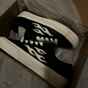 Svarta sneakers med flammor från Straye - Snygga svarta oanvända sneakers från Straye med vita flammor på sidorna. Skorna har en klassisk design med vita snören och en tjock vit sula. Perfekta för en cool och avslappnad stil.