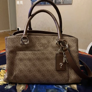 Ljusbrun  axelväska från Guess - Den har flera fickor för att enkelt förvara dina små nödvändigheter och hålla dem alla snygga och rena. Många fickor. Insida: tre stora fack och andra formella fickor inuti. Stängning: dragkedja upptill.