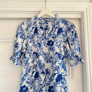 Blåvit blommig klänning från Holly & Whyte - Charmig blåvit blus  från Holly & Whyte med ett vackert blommönster. Blusen har trekvartsärmar och en v-ringad design med volangdetaljer vid halsen. Perfekt för en somrig look!