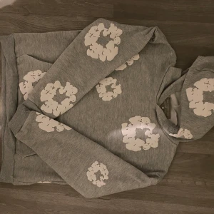 Grå hoodie med blommönster från Denim Tears - Säljer en snygg grå hoodie från Denim Tears med vita blommönster. Tröjan har en klassisk huva och långa ärmar, perfekt för en avslappnad stil. Den är både bekväm och trendig, idealisk för kyligare dagar. Passar som M/L
