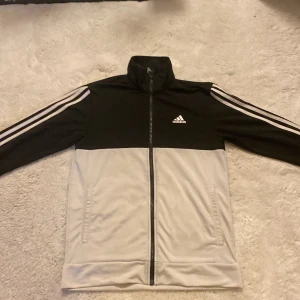Svart och vit jacka från Adidas - Säljer en stilren svart och vit jacka från Adidas med klassiska tre ränder längs ärmarna. Jackan har en dragkedja framtill och Adidas-loggan på bröstet. Perfekt för en sportig look!