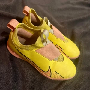 Gula Nike Mercurial fotbollsskor - Säljer ett par gula Nike Mercurial fotbollsskor med rosa detaljer och elastisk snörning. Skorna har en smidig design med Nike Zoom-sula för extra komfort och grepp. Perfekta för inomhusfotboll eller träning.
