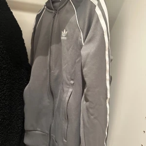 Grå kofta från Adidas - Snygg grå kofta från Adidas med klassiska vita ränder längs ärmarna. Jackan har dragkedja framtill och två fickor. Jag har byxorna till den som matchar bra med den här koftan. Skriv i dm om ni vill ha den här med byxorna så kan vi göra bättre pris. Annars inga fläckar eller skador.