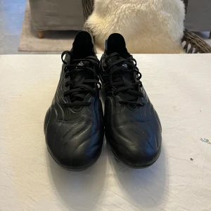 Svarta fotbollsskor från Adidas - Snygga svarta fotbollsskor från Adidas med snörning och dobbar för bra grepp på planen. Skorna har en stilren design med diskreta detaljer i olika färger på hälen.