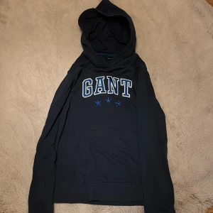 Svart hoodie från GANT - Säljer en stilren svart hoodie från GANT med blå och vita detaljer. Tröjan har en klassisk känguruficka och en bekväm huva. Perfekt för en avslappnad stil. 🖤