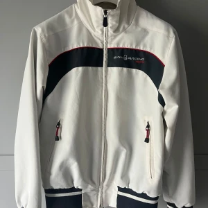 Vit vindjackad från Sail Racing i Gore-Tex - Säljer en stilren vit vindjacka från Sail Racing med svarta och röda detaljer. Jackan har dragkedja framtill och två fickor med dragkedjor. Perfekt för blåsiga dagar med sin höga krage och elastiska muddar.