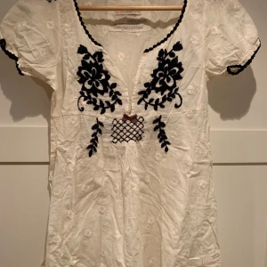 Vit blus från Odd Molly - Charmig vit blus från Odd Molly med broderade blommor i svart och en söt rosett framtill. Klänningen har korta ärmar och en v-ringad design. Perfekt för en somrig look! Färgen är cream vit