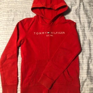 Röd hoodie från Tommy Hilfiger - Säljer en snygg röd hoodie från Tommy Hilfiger med logga på bröstet. Tröjan har en klassisk design med huva och långa ärmar.  Utmärkt skick men säljer pga att den är för liten