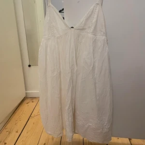 Vit klänning med broderade detaljer - Charmig vit klänning med broderade detaljer och en v-ringad design. Klänningen är ärmlös och har en luftig passform, perfekt för varma dagar. Den har en romantisk känsla med sina fina broderier längs nederkanten.