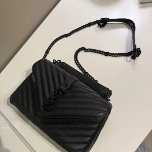 Svart quiltad axelväska INTE DEN RIKTIGA  - Snygg svart axelväska i skinn med quiltad design och kedjerem. Väskan har en elegant logotypdetalj på framsidan och en praktisk storlek för att bära dina nödvändigheter. Perfekt för att ge din outfit en stilfull touch. ””Väskan har dock en liten skada”” Jag har ej använt den men blev skadad av att den ramlade ner från garderoben. 