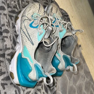 Nike sneakers med turkosa detaljer - Säljer ett par Nike sneakers i beige med turkosa och grå detaljer. Skorna har en modern design med snörning och en bekväm sula. Perfekta för en sportig look.