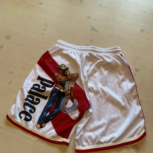 Vita shorts från Palace - Säljer ett par vita shorts från Palace med röd rand och en cool cowboy-grafik på sidan. Shortsen har snörning i midjan och är perfekta för en avslappnad stil. De är gjorda i ett lätt och luftigt material. As skönt silk material på insidan också