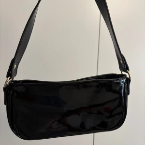 Svart axelväska  - Snygg svart axelväska i glansigt  material. Perfekt för att ge din outfit en stilren touch. Väskan har en enkel design med en justerbar axelrem och dragkedja upptill.