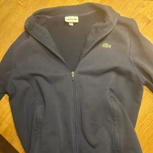 Mörkblå tröja från Lacoste - Säljer en stilren mörkblå tröja från Lacoste med dragkedja och det klassiska krokodilmärket på bröstet. Tröjan har långa ärmar och en bekväm passform. Perfekt för en avslappnad look. Orginal pris på denna hoodien är 1660kr