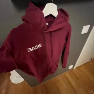 Vinröd hoodie från Base - Säljer en snygg vinröd hoodie från Base med en klassisk design. Hoodien har en justerbar huva med snören och en stor ficka framtill. Perfekt för en avslappnad stil.