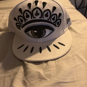 Vit keps från Kenzo - Snygg vit keps från Kenzo med ett unikt ögonmotiv på framsidan. Kepsen har broderad text på baksidan och är en del av New Era 59FIFTY-serien. Perfekt för att ge din outfit en cool touch.