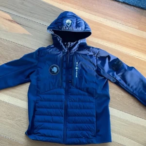 Mörkblå jacka från Sail Racing - Säljer en mörkblå jacka från Sail Racing med huva och dragkedja. Jackan har broderade detaljer och en quiltad design framtill. Perfekt för kyliga dagar med sin stilrena look och funktionella design.