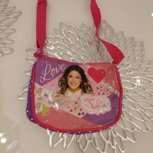 Disney Violetta axelväska - Säljer en färgglad axelväska med motiv från Disney Violetta. Väskan har en justerbar axelrem och ett stort fack med dragkedja. Framsidan är dekorerad med texten 'Love' och ett hjärta. Perfekt för fans av Violetta!