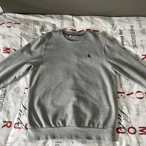 Grå sweatshirt från U.S. Polo Assn. - Säljer en stilren grå sweatshirt från U.S. Polo Assn. med rund hals och ett litet broderat emblem på bröstet. Perfekt för en avslappnad look. Tröjan har långa ärmar och ribbade muddar vid ärmslut och nederkant. Rekommenderad för storlek S mest eftersom sweatshirten är liten för sin storlek. 